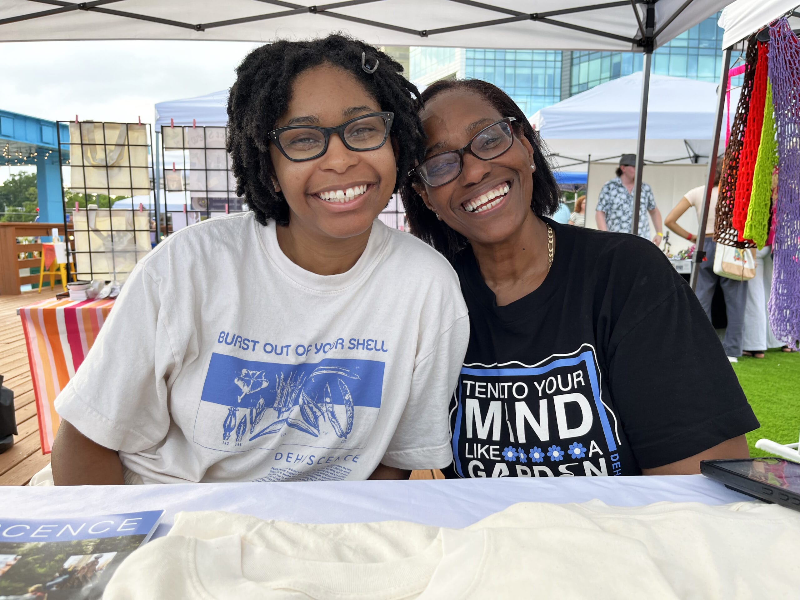 Two people are smiling at a table under a canopy at the night market, where the chill vibe is palpable. One person sports a white t-shirt with a design, while the other wears a black shirt with text. In the background, display booths and art panels complete this lively scene.