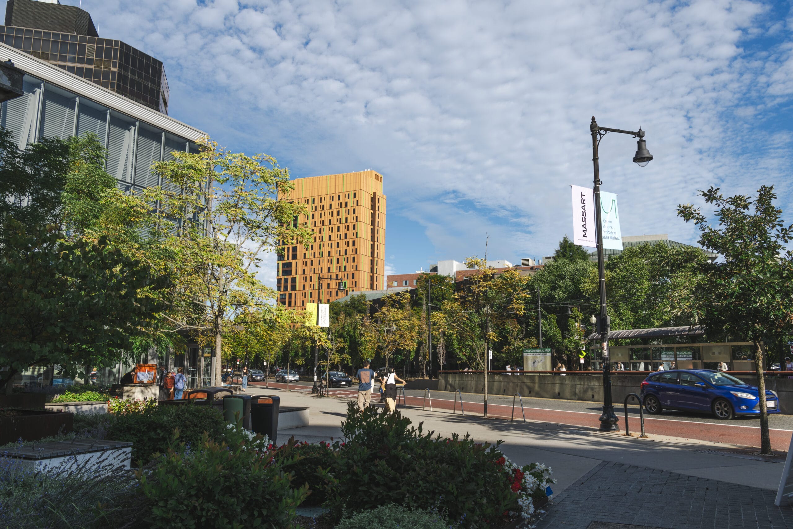 A city street lined with trees and modern buildings, including a distinctive gold-colored tower in the background, where pedestrians walk and cars are parked along the road, reflecting both urban vibrancy and living costs under a partly cloudy sky.