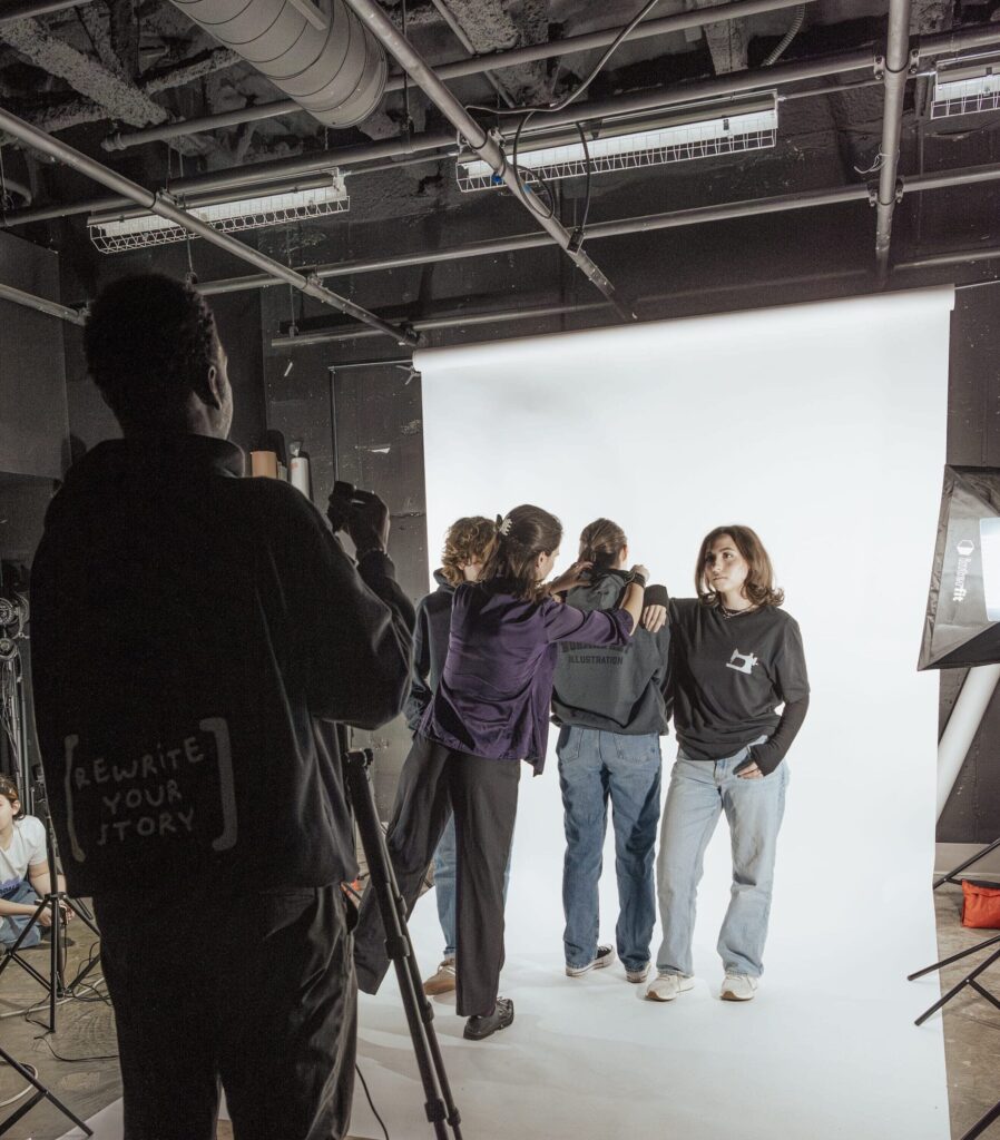 A group of people are setting up for a photo shoot in a studio, collaborating on their creative career. Three pose on a white backdrop while others adjust equipment. One person operates a camera on a tripod in the dimly lit space.